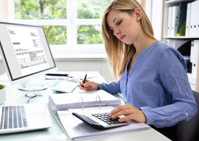 Woman using a calculator and looking at a document while sitting at her computer desk